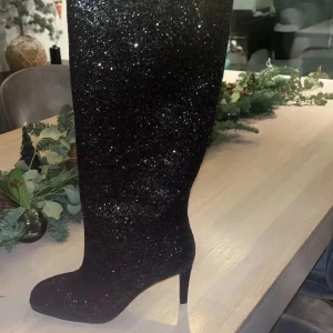 Svarta glittriga stövlar med klack från Nelly - Snygga svarta stövlar med glittrig yta och hög klack. Perfekta för att ge en glamorös touch till din outfit. Stövlarna har en spetsig tå och når upp till knäna. Aldrig använda!
