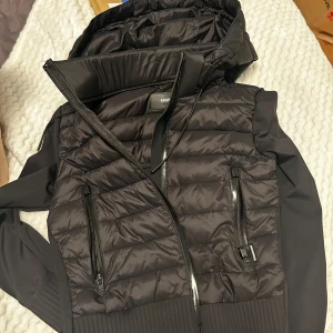 Svart pufferjacka från Khujo - Snygg svart pufferjacka från Khujo med dragkedja och huva. Jackan har långa ärmar och praktiska fickor med dragkedjor. Perfekt för kyligare dagar.