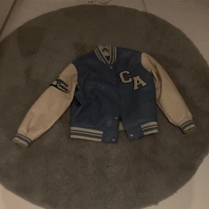 Blå och beige bomberjacka - Snygg bomberjacka i blått och beige med broderade detaljer. Jackan har långa ärmar och knappar framtill. Perfekt för en cool och avslappnad stil.