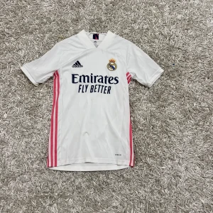 Real Madrid fotbollströja - Säljer en vit Real Madrid fotbollströja från Adidas med korta ärmar. Tröjan har rosa detaljer längs sidorna och klubbens emblem på bröstet. Perfekt för alla fotbollsfans! ⚽