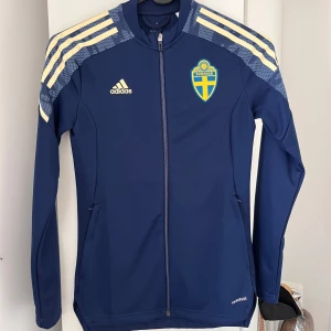 Blå träningsjacka från Adidas storlek XS - Snygg blå träningsjacka från Adidas med Sveriges landslagsemblem på bröstet. Jackan har långa ärmar och dragkedja framtill. Tillverkad av Primeblue-material med Parley Ocean Plastic. Perfekt för sportiga aktiviteter. Använd 1 gång.