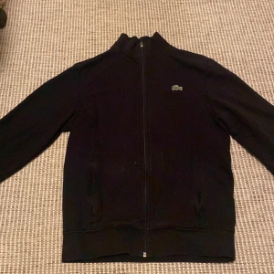 Svart jacka från Lacoste - Säljer en stilren svart jacka från Lacoste med dragkedja och det klassiska krokodilmärket på bröstet. Perfekt för en sportig look.
