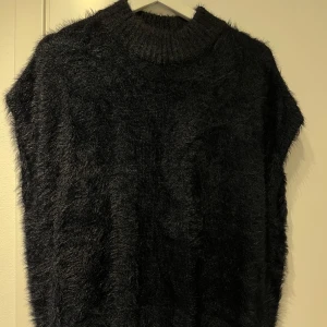 Svart ärmlös stickad tröja - Mysig svart ärmlös stickad tröja med hög krage. Perfekt för lager-på-lager under kyliga dagar. Den har en mjuk och fluffig textur som ger extra värme och stil.