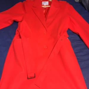 Röd kappa från ONLY - Snygg röd kappa från ONLY med knappar och bälte i midjan. Perfekt för att lysa upp din garderob med en färgstark touch. Kappan har en klassisk design med långa ärmar och en stilren look.