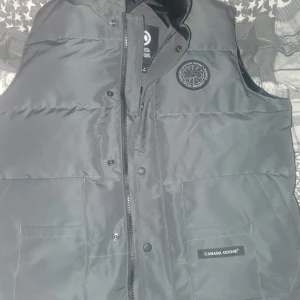 Svart dunväst från Canada Goose - Säljer en svart dunväst från Canada Goose med dragkedja och knappar. Västen har en broderad logotyp på bröstet och är perfekt för kyligare väder.