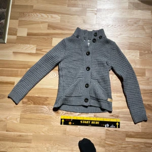 Grå stickad cardigan med knappar - Säljer en snygg grå stickad cardigan med stora svarta knappar. Koftan har en hög krage och ett unikt stickmönster som ger en trendig look. Perfekt för kyliga dagar!