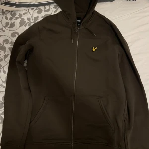 Mörkgrön hoodie från Lyle & Scott - Säljer en snygg brun hoodie från Lyle & Scott med dragkedja och huva. Den har en gul logotyp på bröstet och praktiska fickor framtill. Perfekt för en avslappnad stil. Använd ett par gånger storlek S