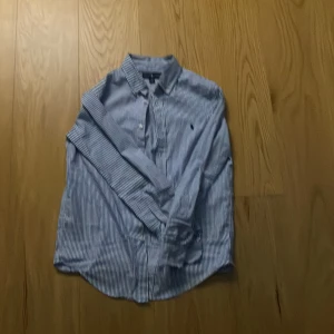 Blå och vit randig skjorta från Ralph Lauren - Snygg blå och vit randig skjorta från Ralph Lauren med klassisk krage och knappar framtill. Skjortan har långa ärmar och det ikoniska Ralph Lauren-logotypen broderad på bröstet. Perfekt för en stilren look.
