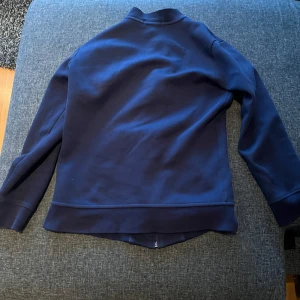 Mörkblå hoodie från Polo Ralph Lauren - Snygg mörkblå hoodie från Polo Ralph Lauren med dragkedja och ribbade muddar. Jackan har långa ärmar och två fickor framtill. Perfekt för en stilren look.