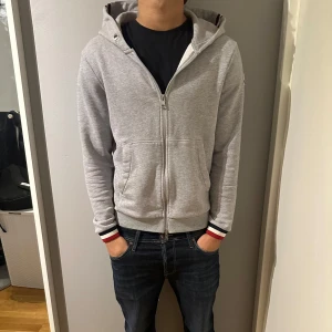 Grå hoodie från Moncler - Tja, säljer en snygg grå hoodie från Moncler med dragkedja och randiga detaljer i rött och vitt på ärmsluten. Perfekt för en avslappnad stil.