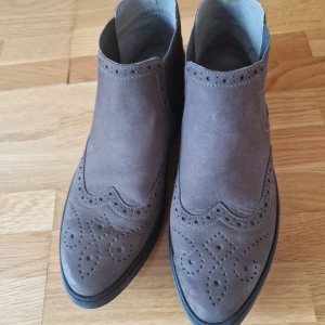 Bruna mockaboots från Ara - Snygga bruna mockaboots från Ara med dekorativa broguedetaljer. Skorna har en robust sula med bra grepp och en dragflik baktill för enkel påtagning. Perfekta för höst och vår