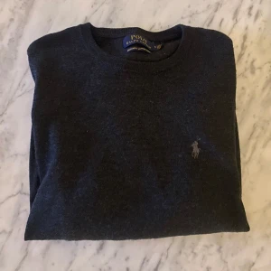 Polo Ralph Lauren tröja - Säljer en stilren mörkgrå tröja från Polo Ralph Lauren i tvättbar merinoull. Tröjan har en klassisk design med rund hals och det ikoniska logotypbroderiet på bröstet. Perfekt för en elegant och bekväm look.