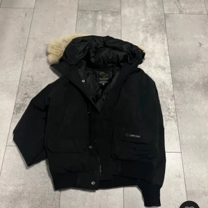 Svart parkajacka från Canada Goose - Säljer en svart parkajacka från Canada Goose med avtagbar päls på huvan. Jackan har dragkedja och knappar framtill samt flera praktiska fickor. Perfekt för kalla vinterdagar. 😃bara höra av er vid frågor😃👊priset är er hugget i stenen