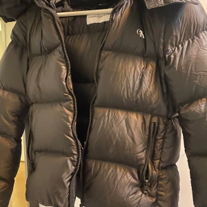 Svart pufferjacka från Calvin Klein Jeans - Snygg svart pufferjacka från Calvin Klein Jeans med dragkedja och huva. Jackan har en stilren design med CK-logga på bröstet och är perfekt för kyligare dagar.