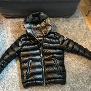 Svart pufferjacka från Colmar - Snygg svart pufferjacka från Colmar med glansig finish och huva. Jackan har dragkedja framtill och två praktiska fickor med dragkedjor. Perfekt för kyliga dagar.