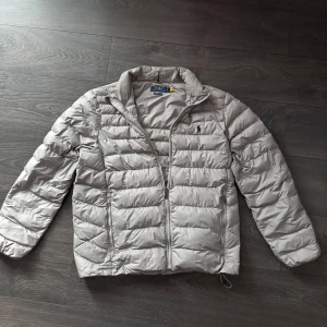 dunjacka från Polo Ralph Lauren - Stilren grå dunjacka från Polo Ralph Lauren med dragkedja framtill som är lätt att matcha. Jackan har en quiltad design och är perfekt för lite kyligare dagar! || Inga defekter/mycket bra skick ||Storlek L. Priset går att diskutera!! Postas eller möts upp i Sthlm. Hör av dig vid ytligare frågor!