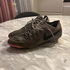 Grå Nike fotbollsskor - Snygga grå Nike fotbollsskor med snörning och svart swoosh-logga. Skorna har en modern design med stickat material och en lätt rosa detalj på sulan. det är det enda som förstör skorna är bara ett litet hål där framme men tror att det inte kan påverka något de är elit bästa modellen av Nike då kostar nya 3500 kolla själv 