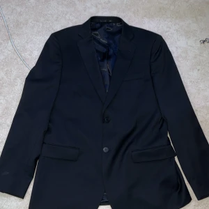 Svart kavaj från Dressmann - Snygg svart kavaj från Dressmann i slim fit. Perfekt för formella tillfällen. Kavajen har en klassisk design med knappar framtill och en elegant insida med röda detaljer.