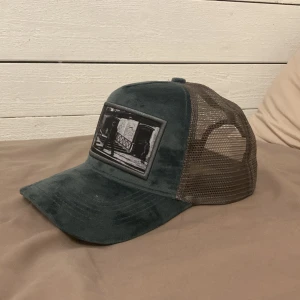 Keps emilio pendeza  - Snygg grön keps med brun nätpanel och justerbar rem baktill. Framtill finns en svartvit broderad patch med ett motiv. Perfekt för en avslappnad stil.