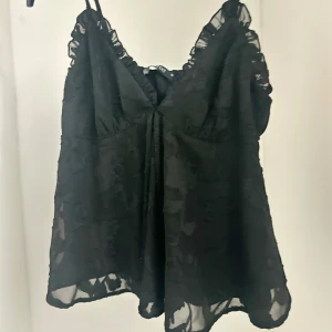 Svart blommig linne - Snygg svart linne med blommigt mönster och volangdetaljer vid axelbanden. 
