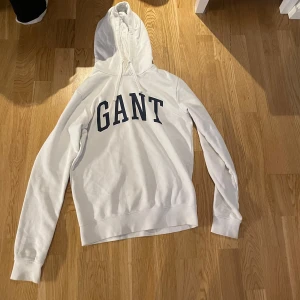 Vit hoodie från GANT - Säljer en stilren vit hoodie från GANT med stor logotyp i marinblått på framsidan. Tröjan har en klassisk design med huva och långa ärmar.