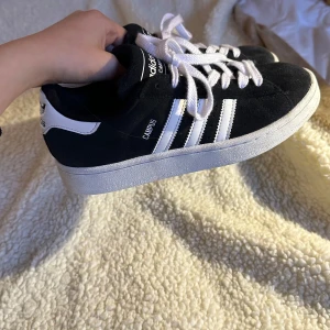 Svarta Adidas Campus sneakers - Säljer ett par klassiska svarta Adidas Campus sneakers med vita detaljer och snörning. Skorna har den ikoniska tre-randiga designen på sidorna och en bekväm vit sula. Perfekta för en avslappnad stil. De blev för små❤️