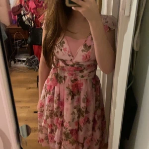 Rosa blommig klänning - Charmig rosa klänning med blommigt mönster och omlott-design. Den har korta ärmar och en v-ringad hals. Perfekt för sommarens festligheter. ❤️storlek M men passar även xs och s man kan välja hur hårt man väljer att knyta den❤️