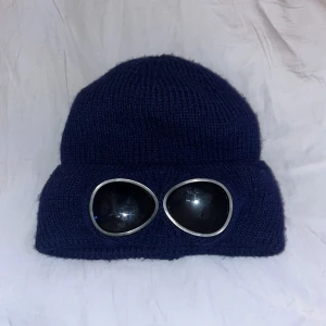 Mörkblå stickad mössa med glasögondetalj - Snygg mörkblå stickad mössa med unika glasögondetaljer framtill. Insidan är fodrad med mjukt, grått material för extra värme. Perfekt för kalla dagar.