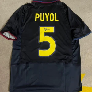 Svart fotbollströja med Puyol 5 - Säljer en svart fotbollströja med Puyol och nummer 5 på ryggen. Tröjan har korta ärmar och detaljer i rött och blått. Perfekt för fotbollsfans!