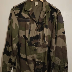 Camouflage militärjacka med nitar - Säljer en cool militärjacka i camouflage-mönster med nitar på axlarna. Jackan har långa ärmar och flera fickor framtill. Perfekt för en edgy look!