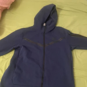 Blå huvtröja från Nike - Säljer en blå huvtröja från Nike med dragkedja framtill. Tröjan har en huva och långa ärmar, perfekt för en avslappnad stil. Den är tillverkad i ett mjukt material som känns skönt mot huden. Har knappast haft den köpte får en månad sen tyckte den var för stor 