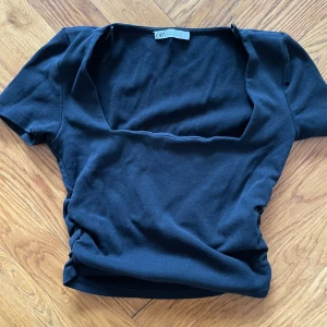 Svart topp från Zara - Säljer en stilren svart topp från Zara med fyrkantig urringning. Toppen har korta ärmar och är tillverkad i ett mjukt material som ger en bekväm passform. Croppad passform och detaljer i sidorna.