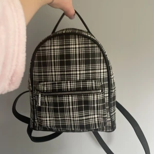 Svart och vit rutig ryggsäck - Snygg och praktisk ryggsäck med svart och vitt rutigt mönster. Väskan har en dragkedja framtill och justerbara axelremmar. Perfekt för att bära dina dagliga nödvändigheter med stil.