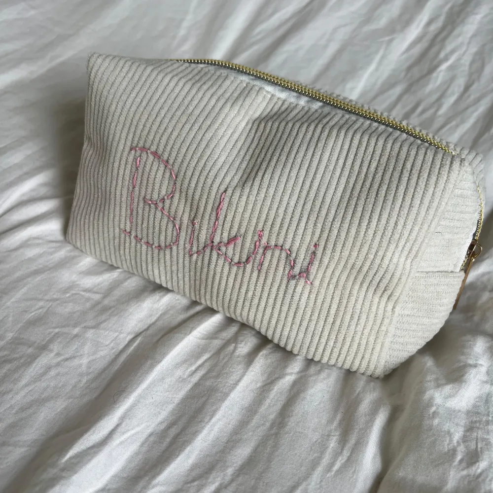 En vit ribbad förvaringspåse med texten 'Bikini' (går även att be om annan personlig broderad text) broderad i rosa. Smidig att ta med på resan! Storlek: 17x10x6cm. Laukut & Käsilaukut.