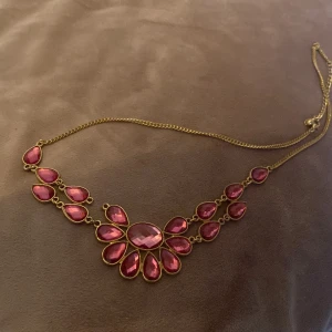 Guldhalsband med rosa stenar - Elegant guldhalsband med stora rosa stenar i en blommig design. Kedjan är tunn och smidig, vilket ger en fin kontrast till de iögonfallande stenarna. Perfekt för att ge en färgklick till din outfit.