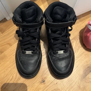 Svarta Nike Air Force sneakers - Säljer ett par klassiska svarta Nike Air Force sneakers med snörning och hög ankelrem. Skorna har en perforerad tå för ventilation och en robust sula för bra grepp. Perfekta för en stilren look. I bra använt skick. 