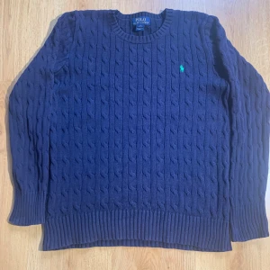 Mörkblå stickad tröja från Ralph Lauren - Säljer en snygg mörkblå stickad tröja från Ralph Lauren med kabelstickat mönster och det klassiska gröna logotypbroderiet på bröstet. Tröjan har långa ärmar och ribbstickade muddar vid ärmslut och nederkant. Perfekt för kyliga dagar! Passar dig som är 164