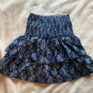 Blå volangkjol med mönster - Charmig blå kortkjol med vitt mönster och volanger. Kjolen har en elastisk midja för bekväm passform och är perfekt för sommarens äventyr. Den är gjord i ett lätt och luftigt material som ger en härlig känsla. Har insydda shorts. Aldrig använd så skicket är 10 av 10. Vid frågor skicka dm.