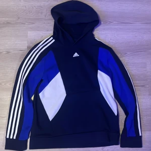 Blå och vit hoodie från Adidas - Säljer en snygg blå hoodie från Adidas med vita och blå detaljer. Den har de klassiska tre ränderna längs ärmarna och Adidas-loggan på bröstet. Perfekt för en sportig look eller avslappnad stil. 🏃‍♂️