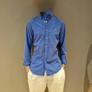 Blå skjorta från Ralph Lauren - Snygg blå skjorta från Ralph Lauren med klassisk krage och knappar framtill. Skjortan har långa ärmar och en liten broderad logga på bröstet. Perfekt för en stilren look. Passar M/S