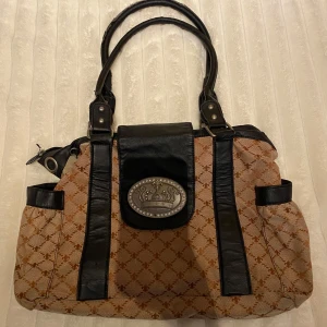 Mönstrad handväska med svarta detaljer - Säljer en snygg handväska med brunt mönster och svarta detaljer. Väskan har två handtag och en dekorativ metallplatta framtill.  Väskan är i väldigt fin kvalitet förutom att handtaget har lite mindre skador. Samt att metallplattan saknar en diamant  Skriv gärna för fler bilder!💋