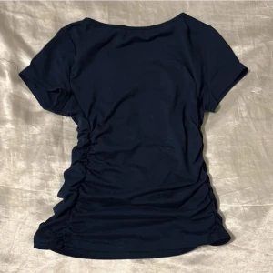 Mörkblå kortärmad topp - Säljer en stilren mörkblå topp med korta ärmar. Toppen har en rynkad detalj på sidorna som ger en fin passform. Perfekt för en avslappnad look.