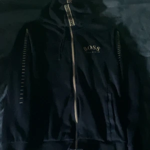 Svart tracksuit  från Hugo Boss - Snygg svart hoodie från Hugo Boss med dragkedja och stilrena detaljer längs ärmarna. Perfekt för en casual look med en touch av lyx. Märkesloggan är tydligt synlig på bröstet.