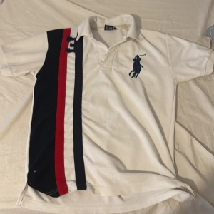 Vit pikétröja med ränder från Ralph Lauren - Säljer en klassisk vit pikétröja från Ralph Lauren med korta ärmar. Tröjan har en stor logotyp på bröstet och dekorativa ränder i svart och rött på ena sidan. Perfekt för en stilren och sportig look.