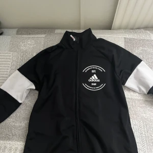 Kofta från Adidas - Snygg svart  kofta från Adidas med dragkedja framtill. Jackan har långa ärmar och ett tryck på bröstet med Adidas-loggan och text. Perfekt för en sportig look! Storlek M