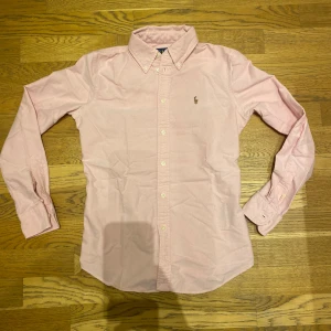 Rosa skjorta från Ralph Lauren - Snygg rosa skjorta från Ralph Lauren i storlek S. Skjortan har en klassisk krage och är dekorerad med det ikoniska broderade logotypen på bröstet. Perfekt för en stilren look.