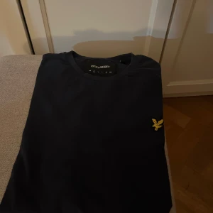 Svart t-shirt från Lyle & Scott - Snygg svart t-shirt från Lyle & Scott med deras ikoniska gula logga på bröstet. Perfekt för en stilren och avslappnad look. Passar bra till jeans eller shorts. Skick 9/10