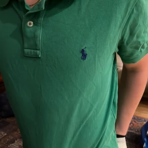 Grön pikétröja från Ralph Lauren - Säljer en klassisk grön pikétröja från Ralph Lauren med den ikoniska loggan broderad i blått på bröstet. Tröjan har en knappslå med två knappar och korta ärmar. Perfekt för en avslappnad stil. Nypris ca 800kr kommer tvättad och struken