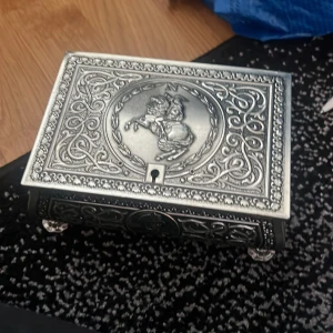 Dekorativ smyckeskrin i silverfärg - Elegant smyckeskrin i silverfärg med detaljerade mönster och en ryttare på locket. Insidan är klädd i röd sammet, vilket ger en lyxig känsla. Perfekt för att förvara dina smycken eller små skatter.