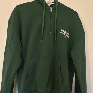 Grön hoodie  - Säljer en grön hoodie från Wardrobe Essentials med dragkedja och känguruficka. Perfekt för en avslappnad stil. Den har en justerbar huva med snören och ribbade muddar vid ärmslut och nederkant.